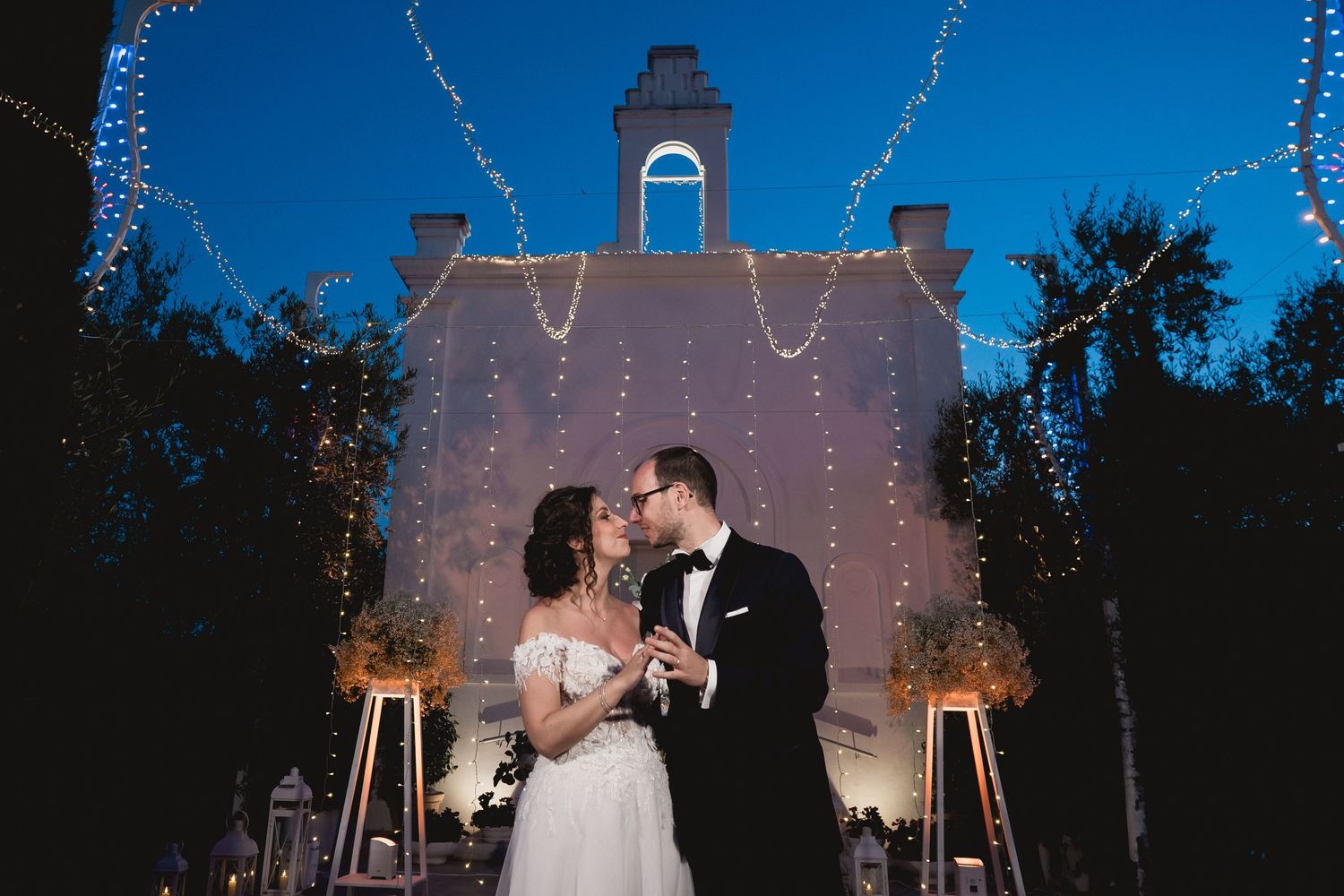 Couple sharing intimate moment in front of illuminated chapel with string lights at dusk.