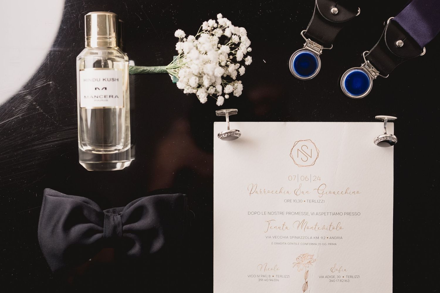 Wedding details flatlay with bowtie, invitation, cologne bottle and white flowers on dark surface.