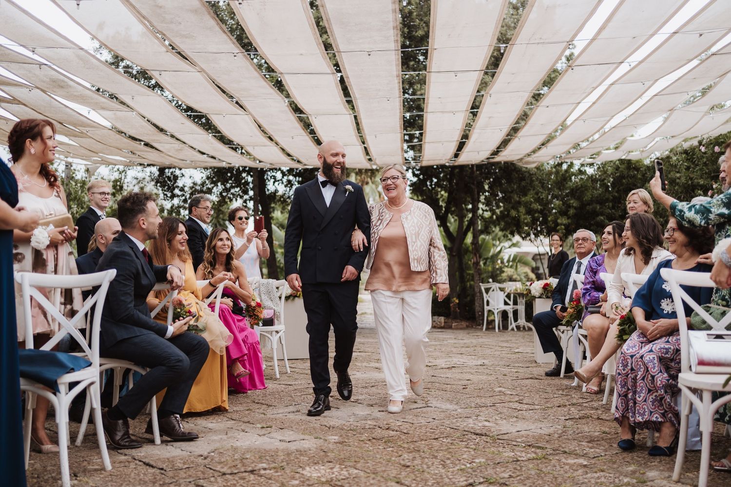 Wedding ceremony under a draped canopy with guests seated along aisle during procession.