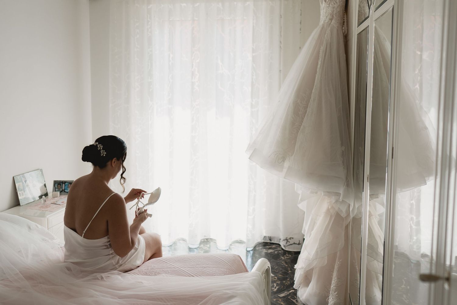 Bride sitting on bed in white slip dress looking at wedding gown hanging nearby.