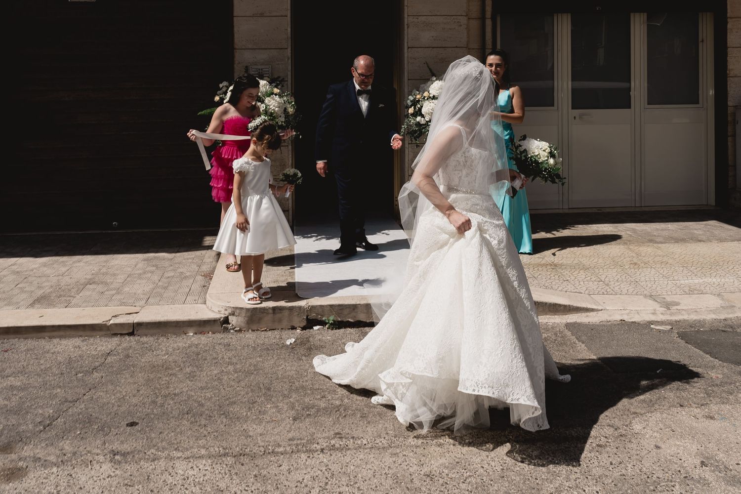 Wedding party exits church in bright sunlight with flowing white dress in motion.
