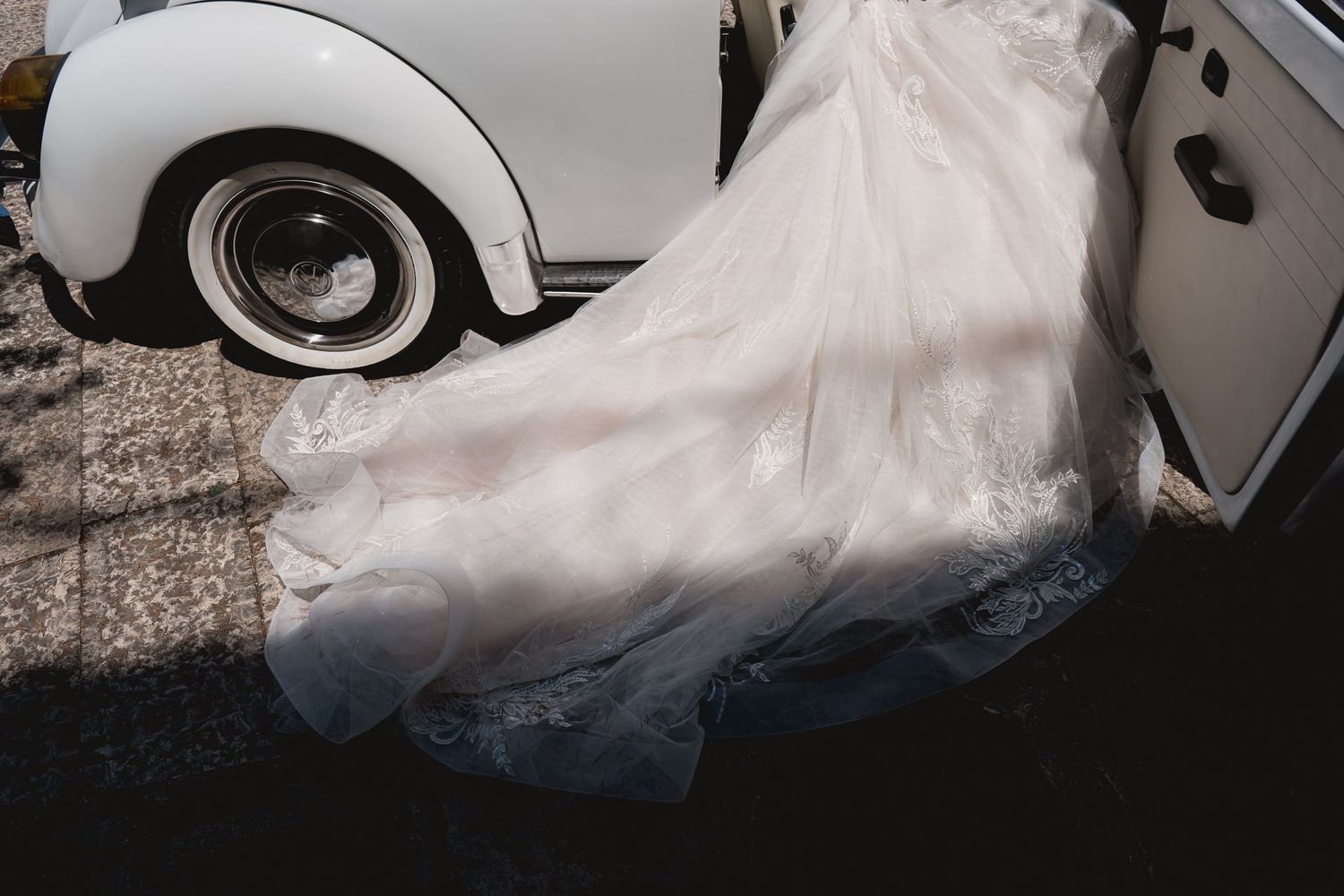 The flowing train of a white wedding dress beside a classic vintage car wheel.