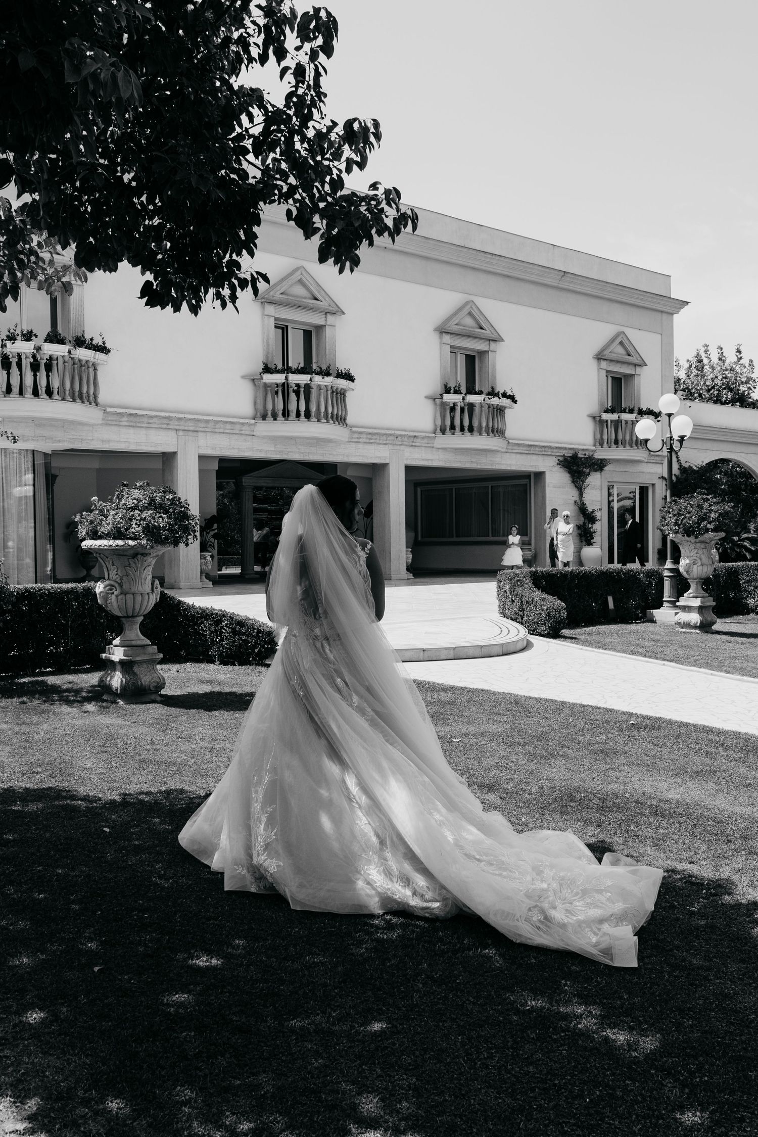 A bride in a flowing wedding dress and veil stands before a classic white mansion.