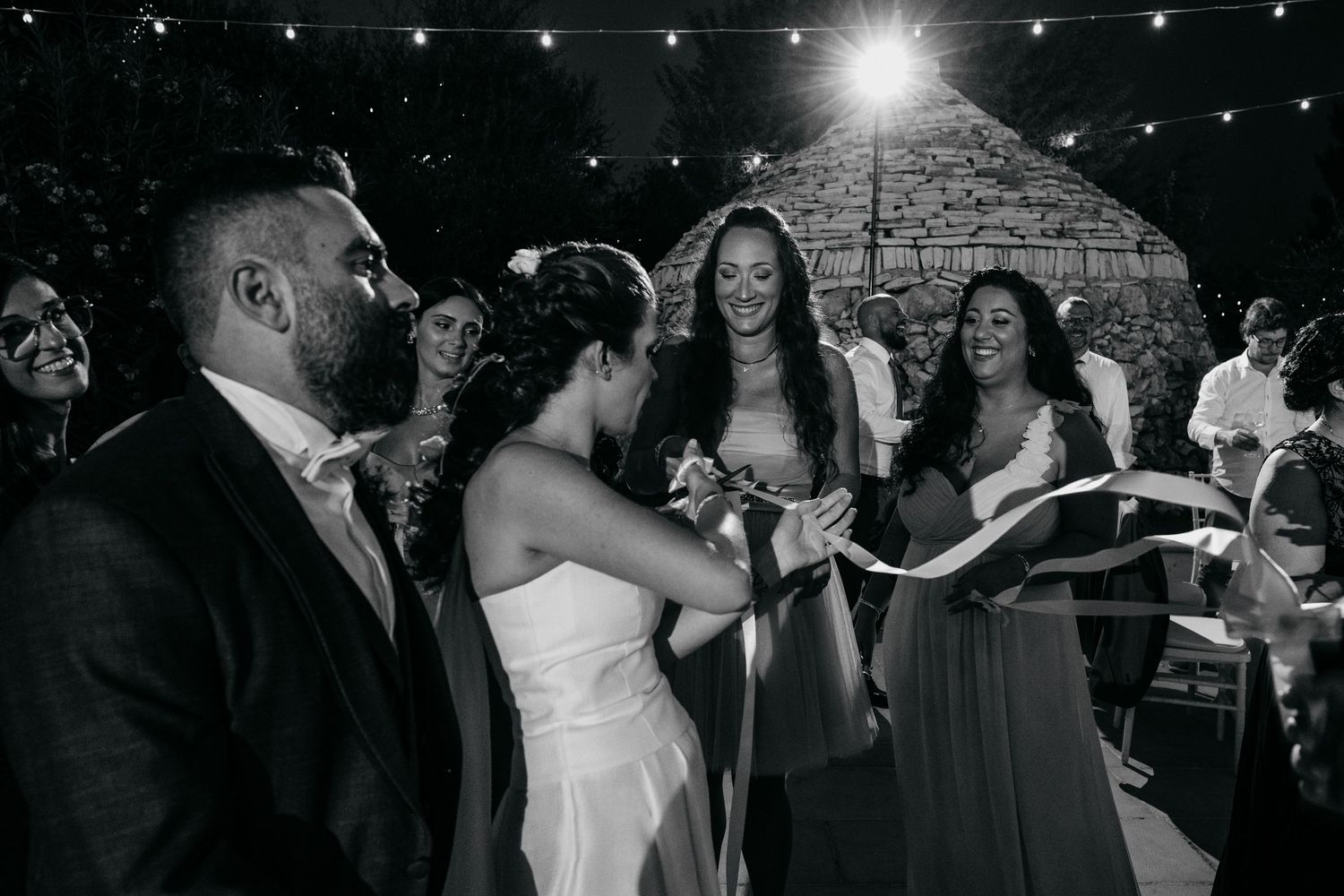 Wedding party celebrates together under string lights at a nighttime outdoor reception.
