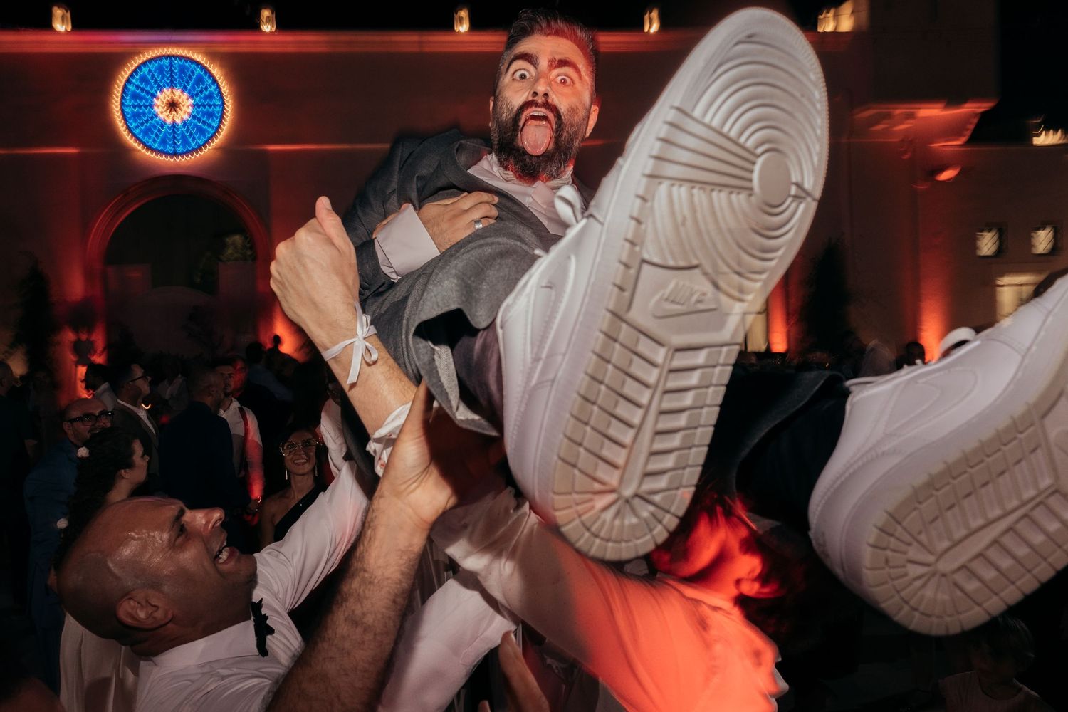 Dynamic party scene shows people lifting and celebrating with visible sneakers in red-lit atmosphere.