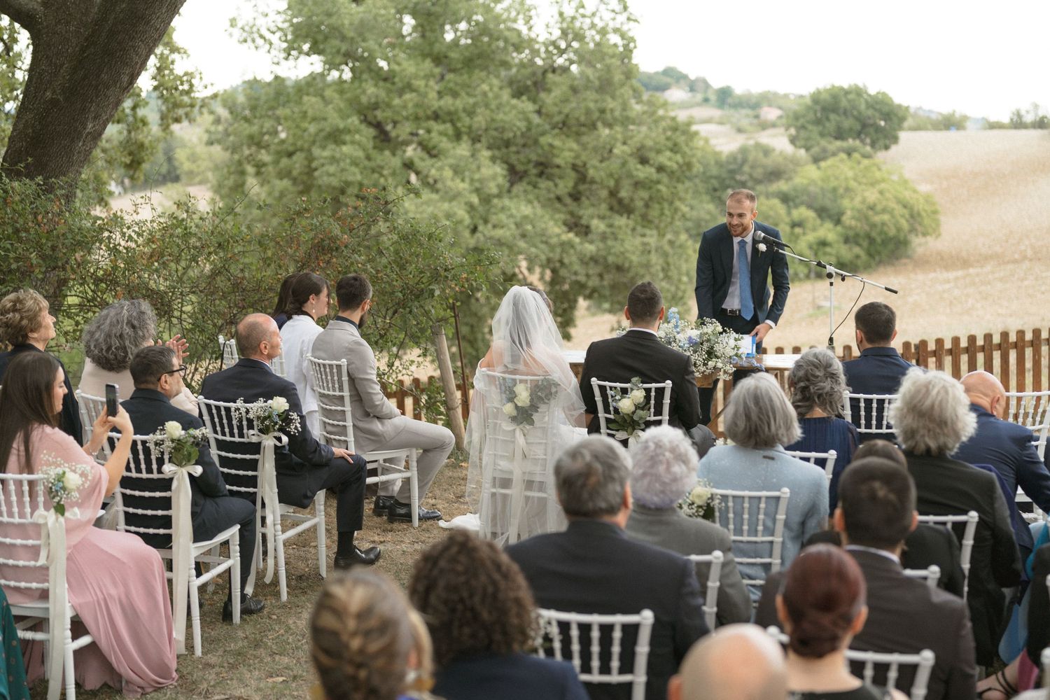 Outdoor wedding ceremony setup with white chairs under a tree in natural lighting.