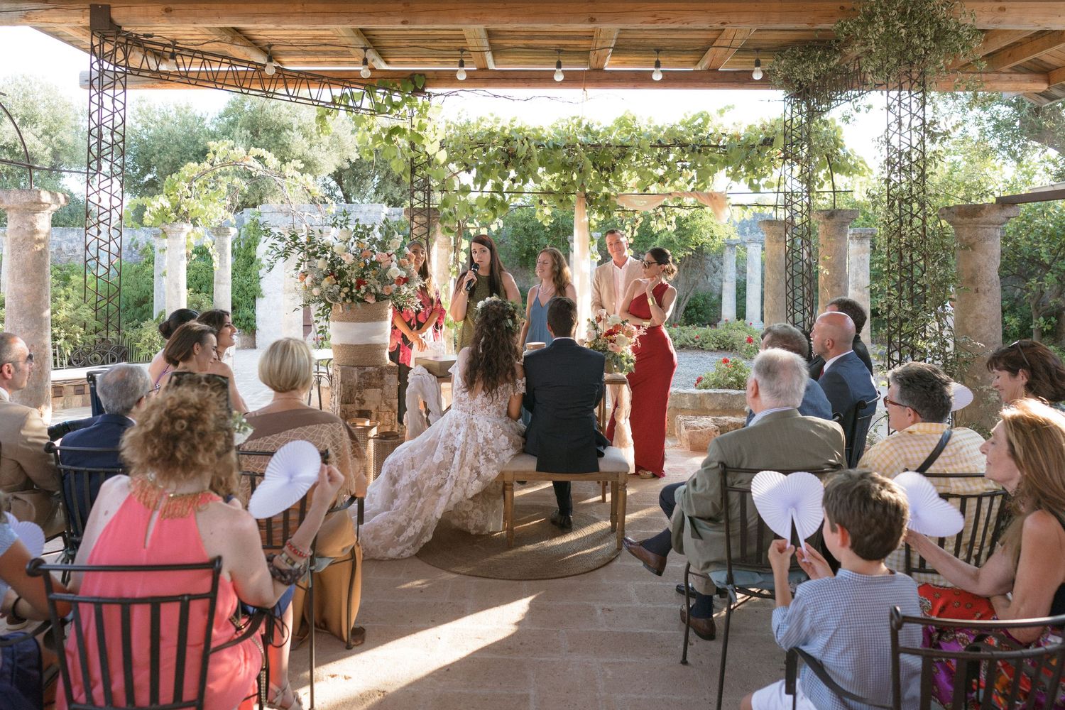 Wedding ceremony taking place under a rustic wooden pergola with guests seated in white chairs.