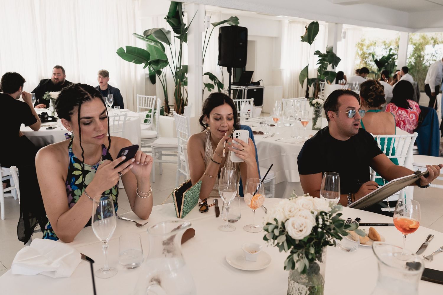 Wedding guests enjoy dinner at an elegant reception table decorated with tropical greenery and white linens.