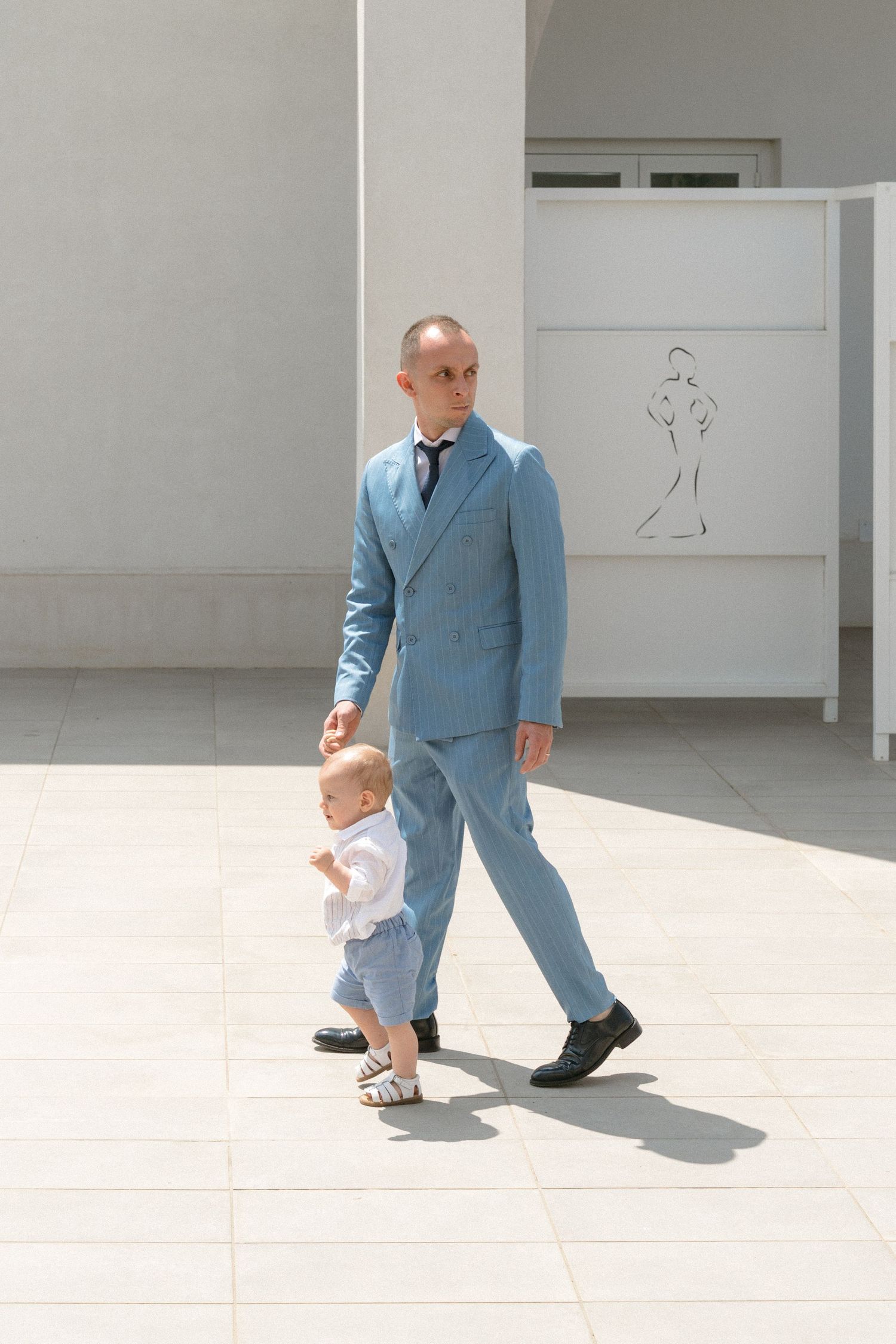 Father and young child walking together in matching light blue suits in a bright minimalist space.