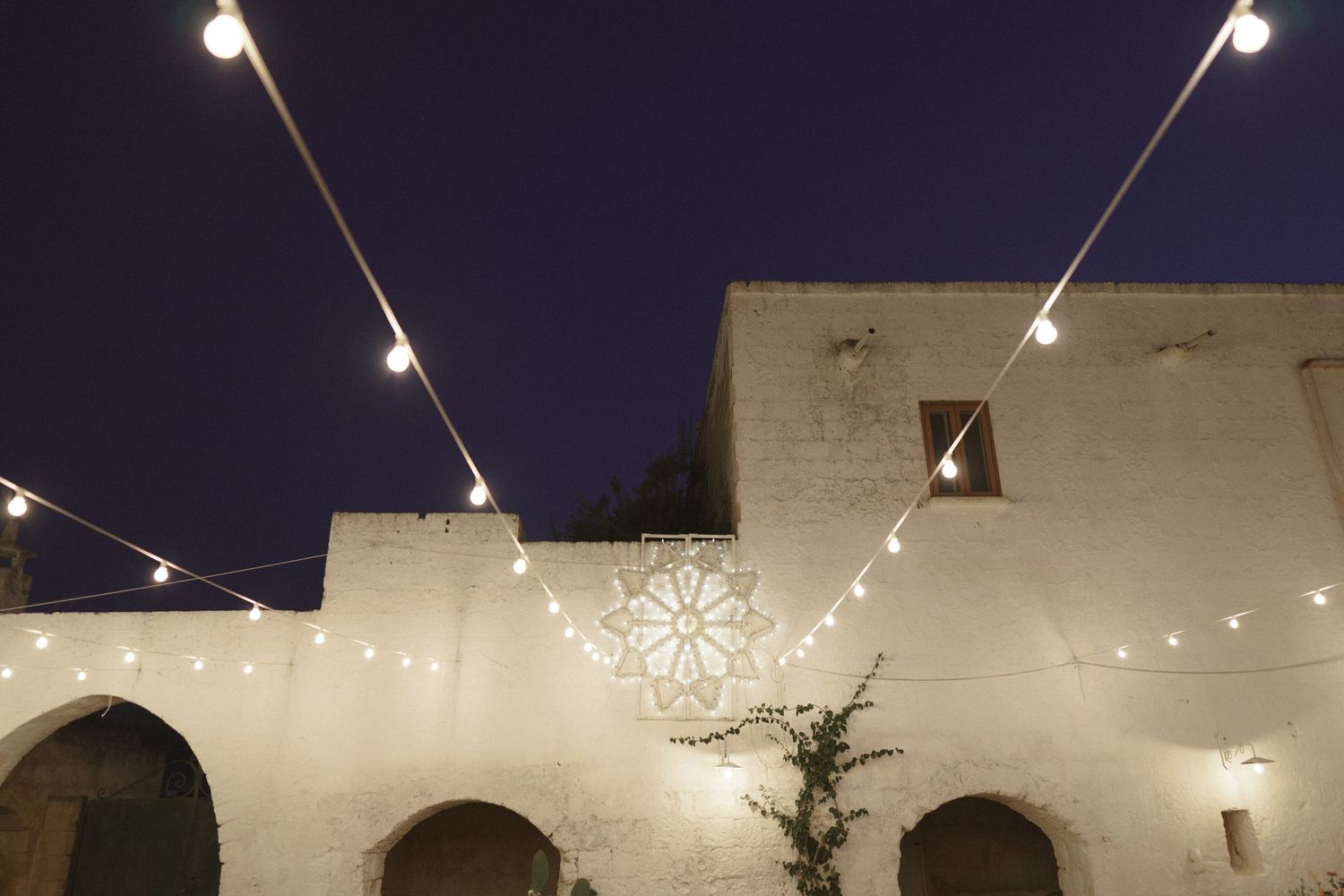 String lights illuminate an outdoor courtyard with white arches against a dark night sky, creating a magical atmosphere.