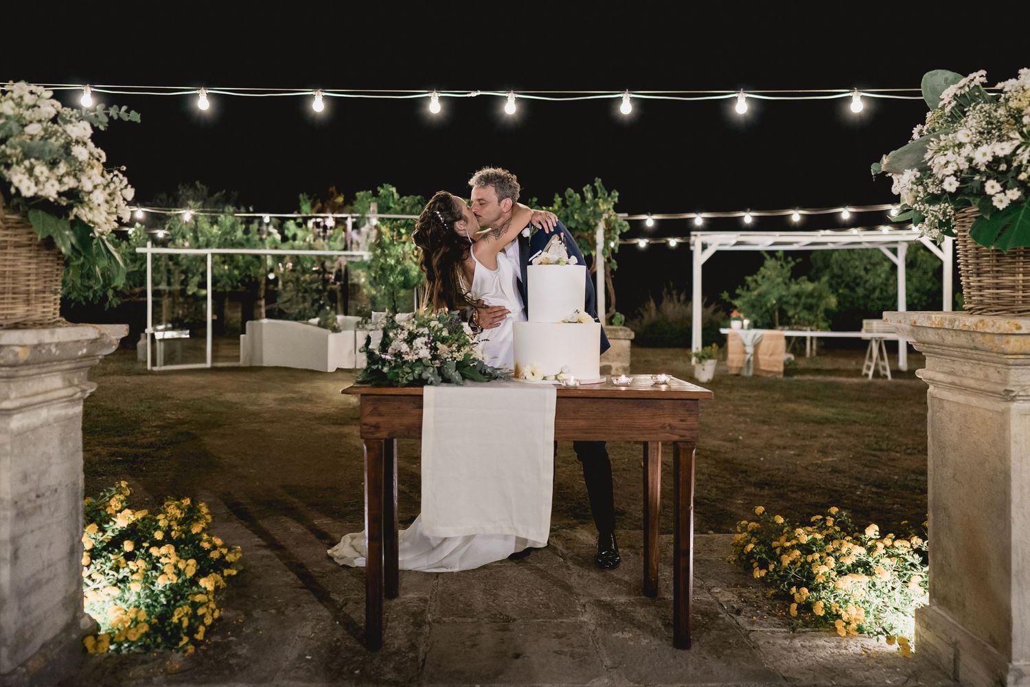Romantic outdoor evening wedding ceremony with string lights overhead and beautiful floral arrangements on rustic table.