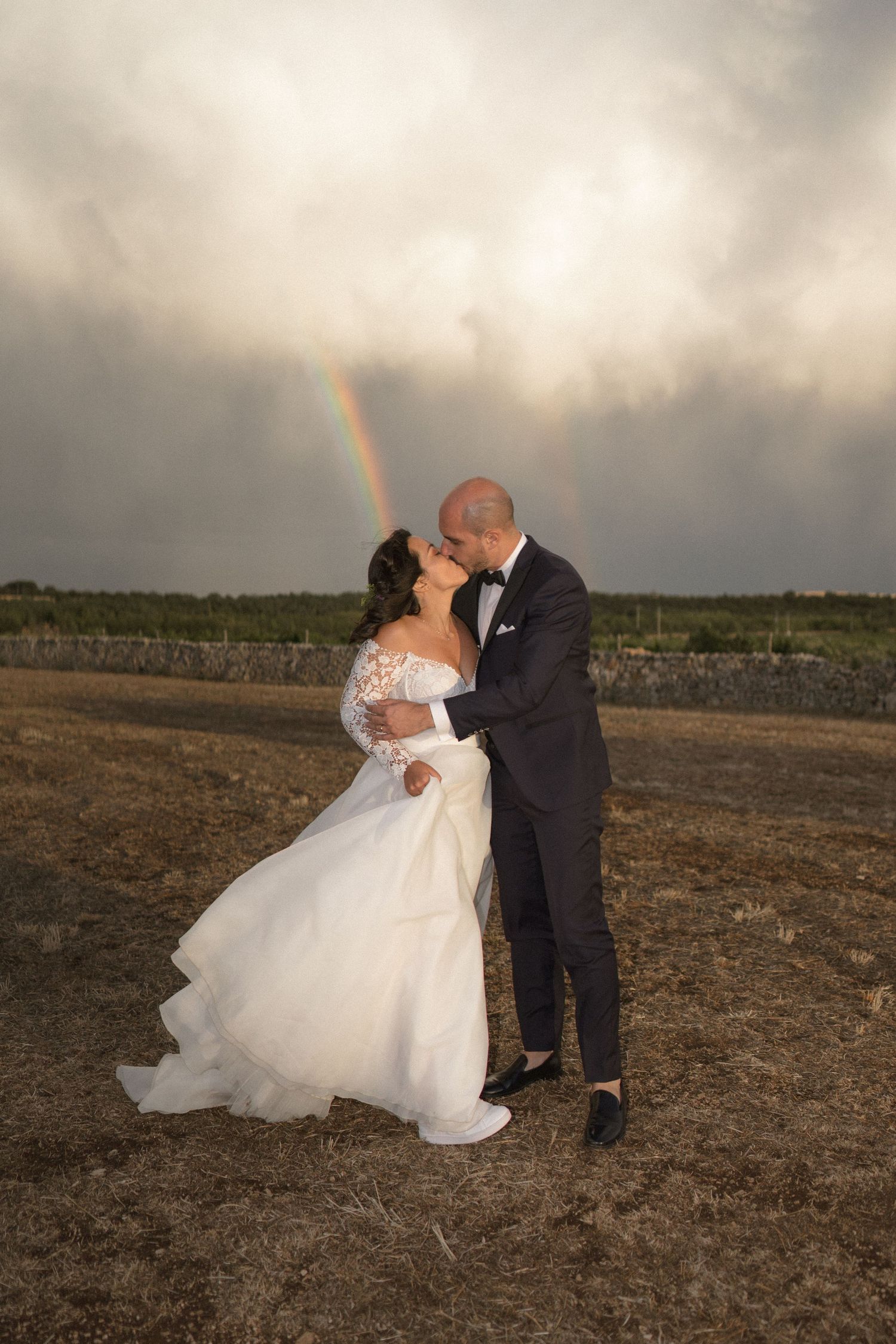 Wedding couple dancing in a field at sunset with a rainbow appearing in the cloudy sky.