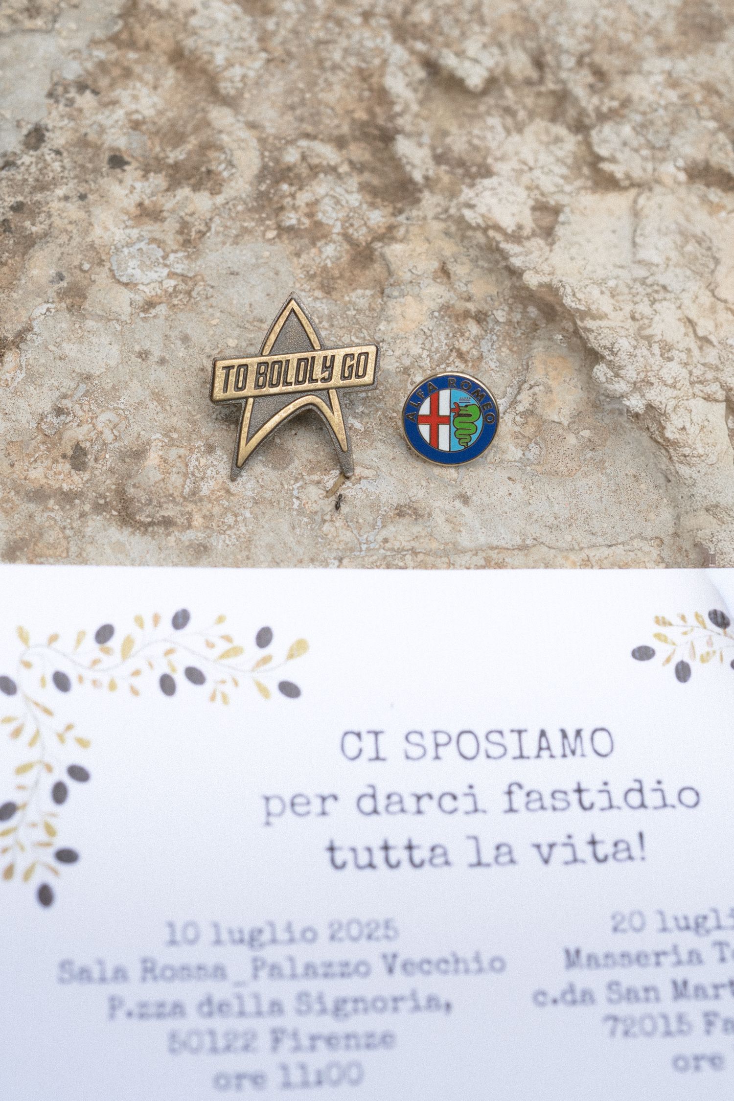 Wedding invitation details and decorative pins displayed on textured beige stone surface with Italian text.