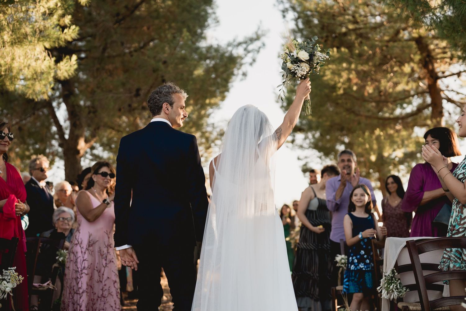 A wedding couple celebrates with raised arms as guests cheer during an outdoor ceremony.