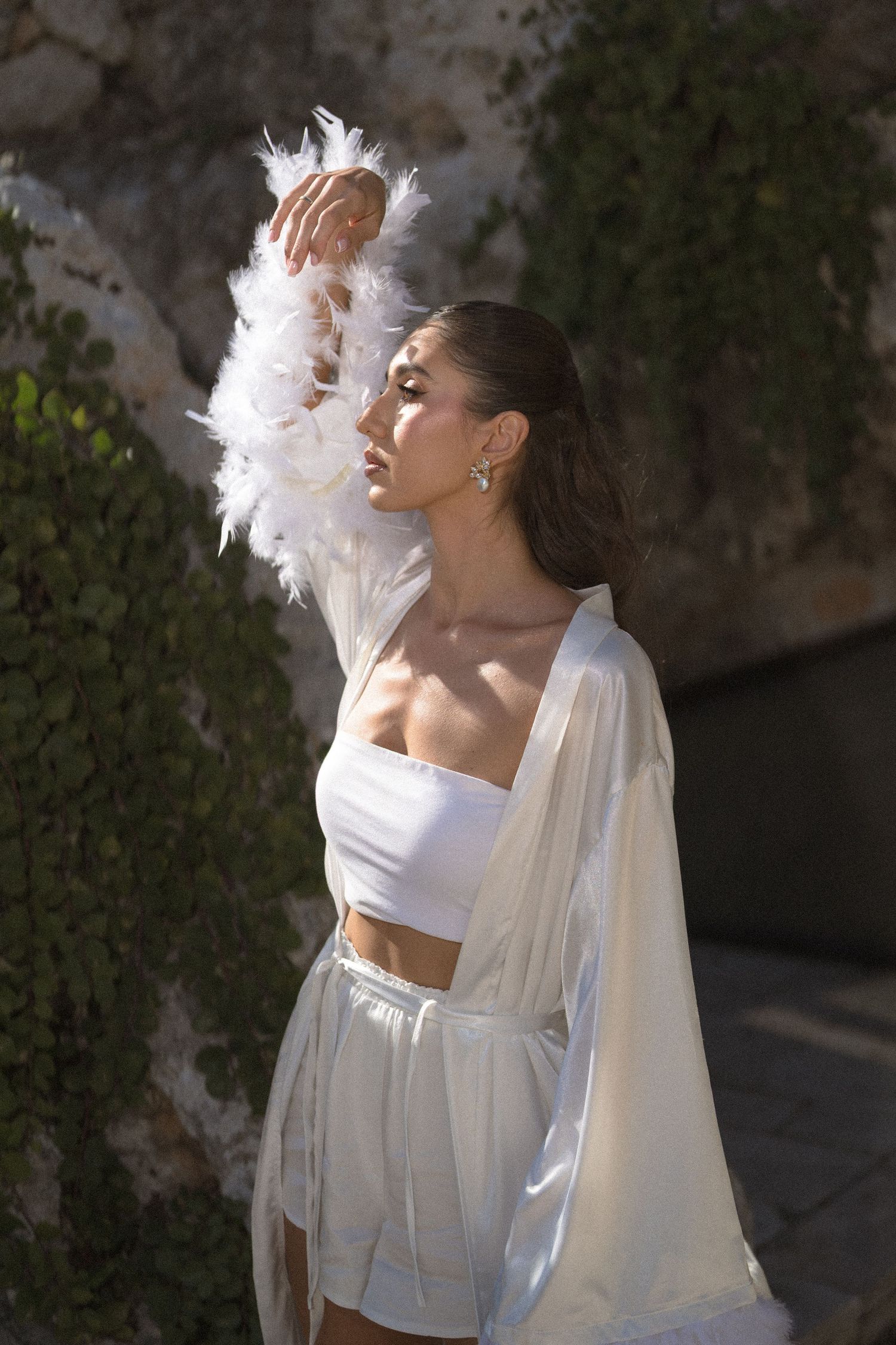 A woman in a white satin robe with feathered sleeves raises her arm, standing before an ivy-covered stone wall in sunlight.