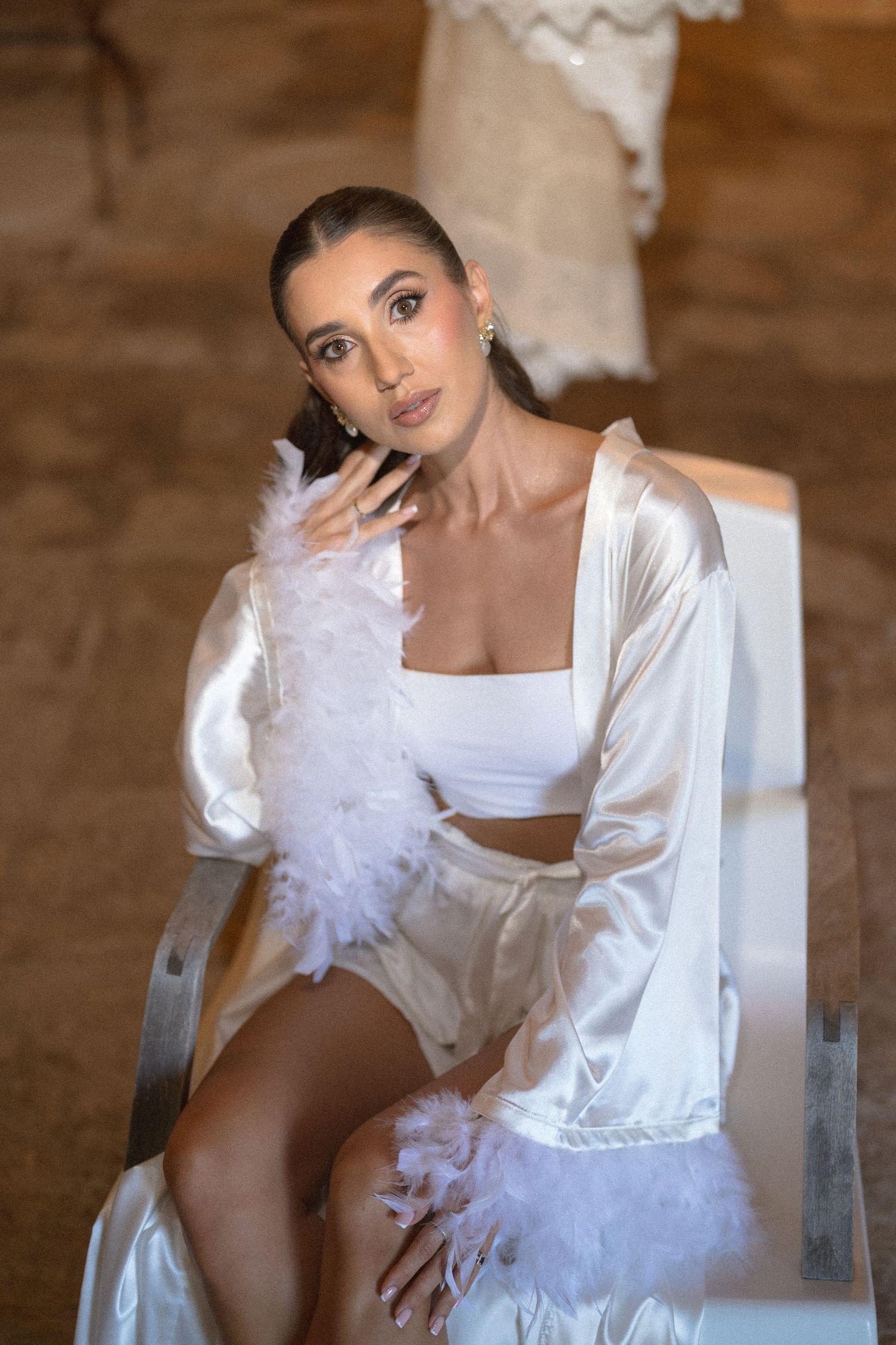 Woman in a white satin robe and crop top with feather-trimmed sleeves sits on a chair, posing for a bridal boudoir portrait.