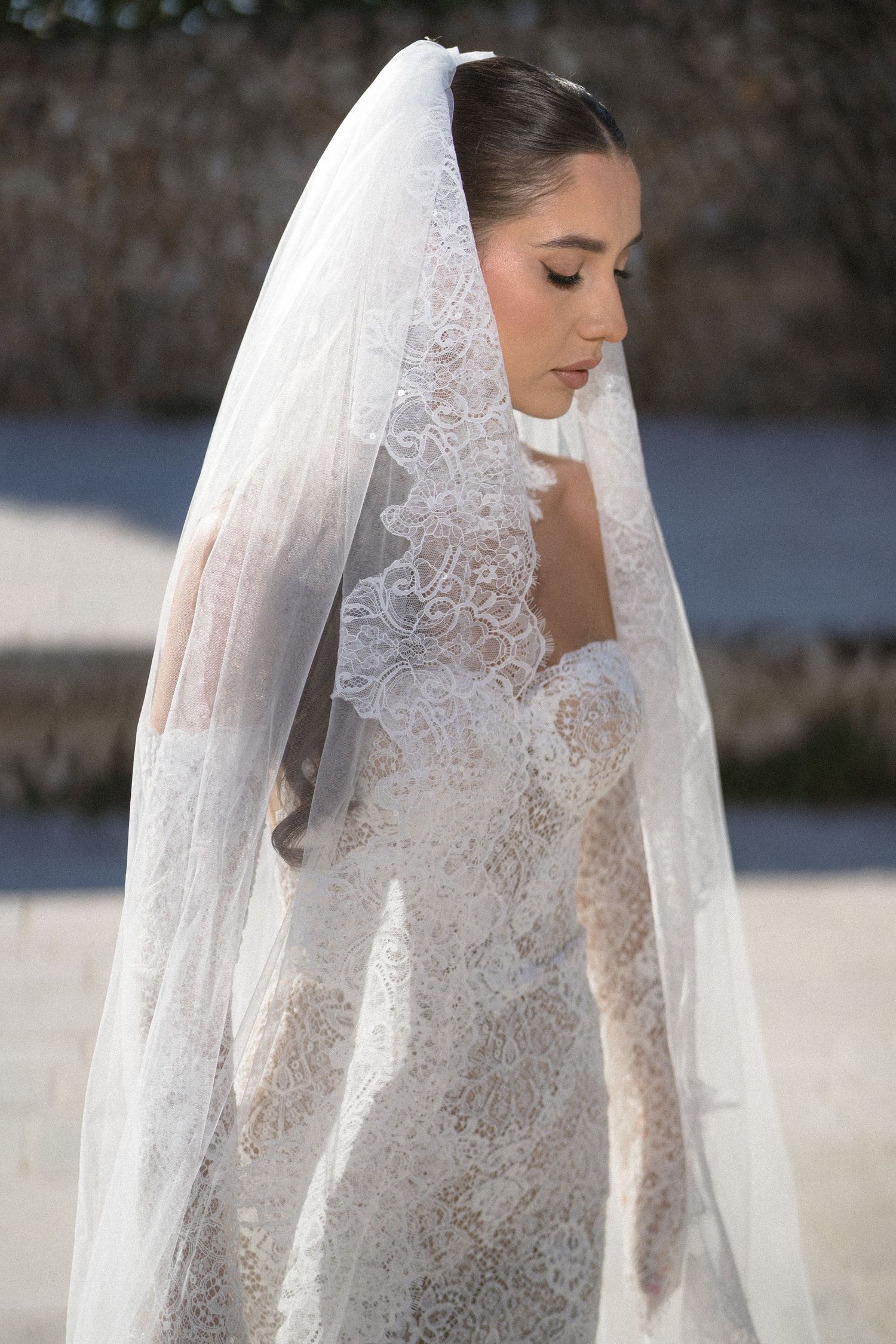 Side profile of a bride in a strapless lace wedding dress and a long lace-edged veil, outdoors in soft daylight.