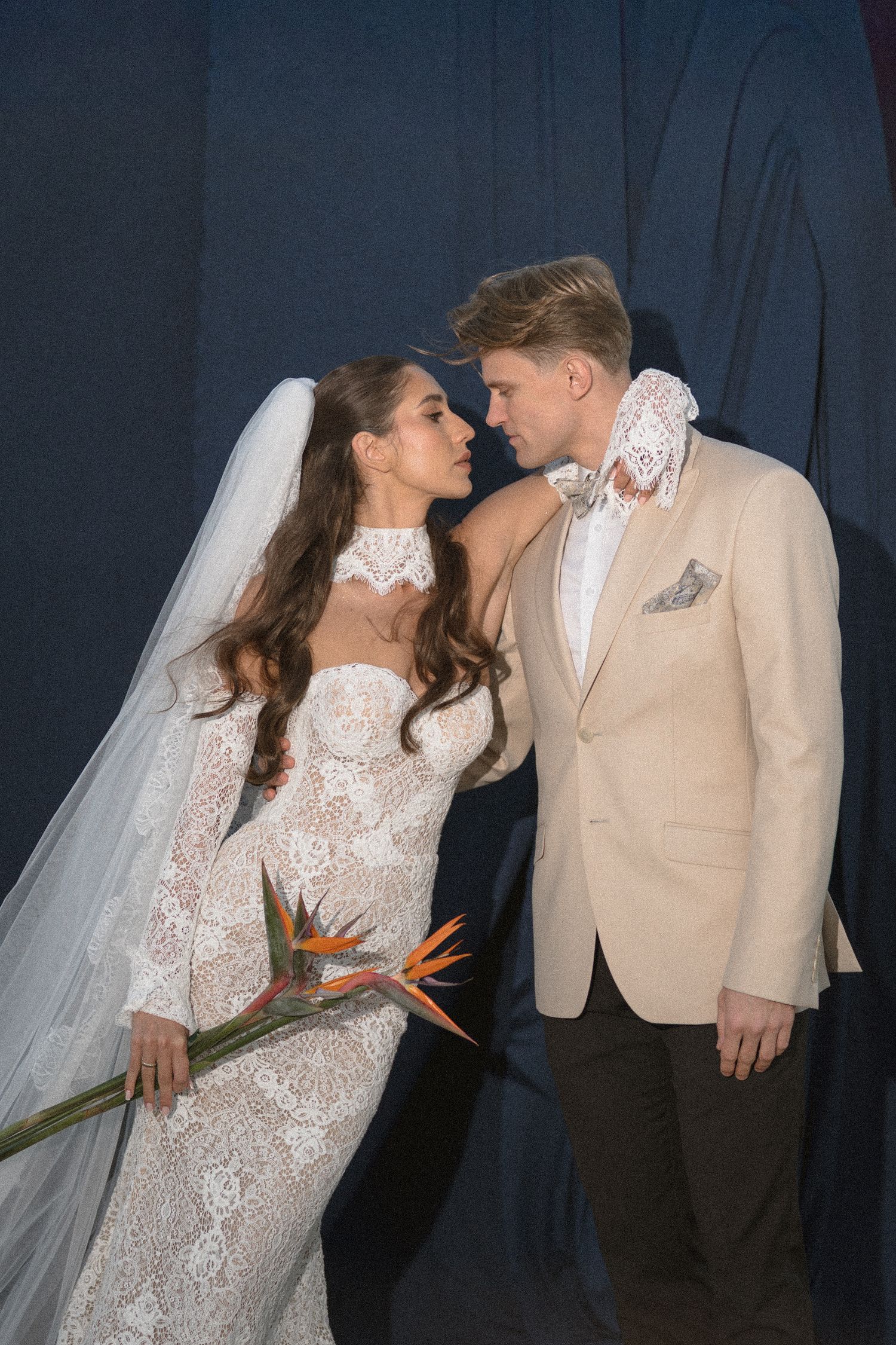 Bride in a lace gown holding a bird of paradise flower leans toward her groom in a beige blazer against a dark blue backdrop.