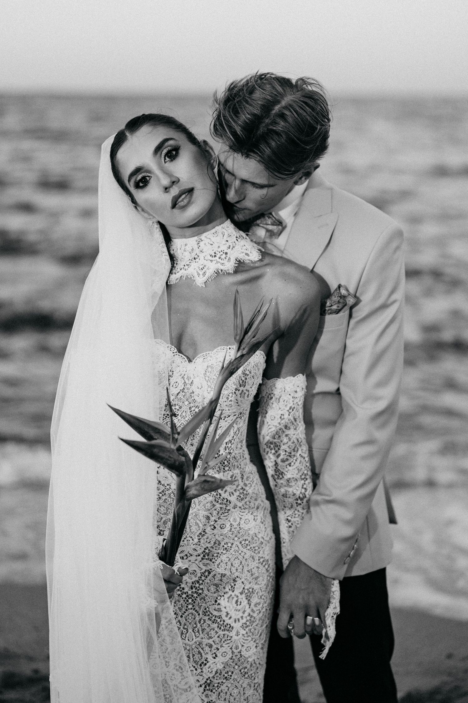 Black and white beach wedding portrait of a groom kissing his bride's shoulder as she holds a flower in a lace gown and veil.