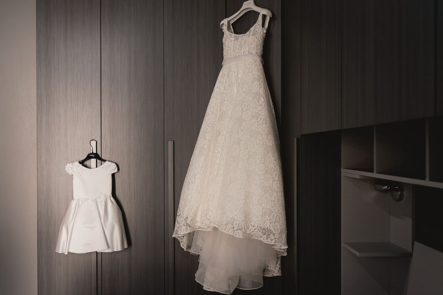 Wedding dress and flower girl dress hanging side by side in a dimly lit room.