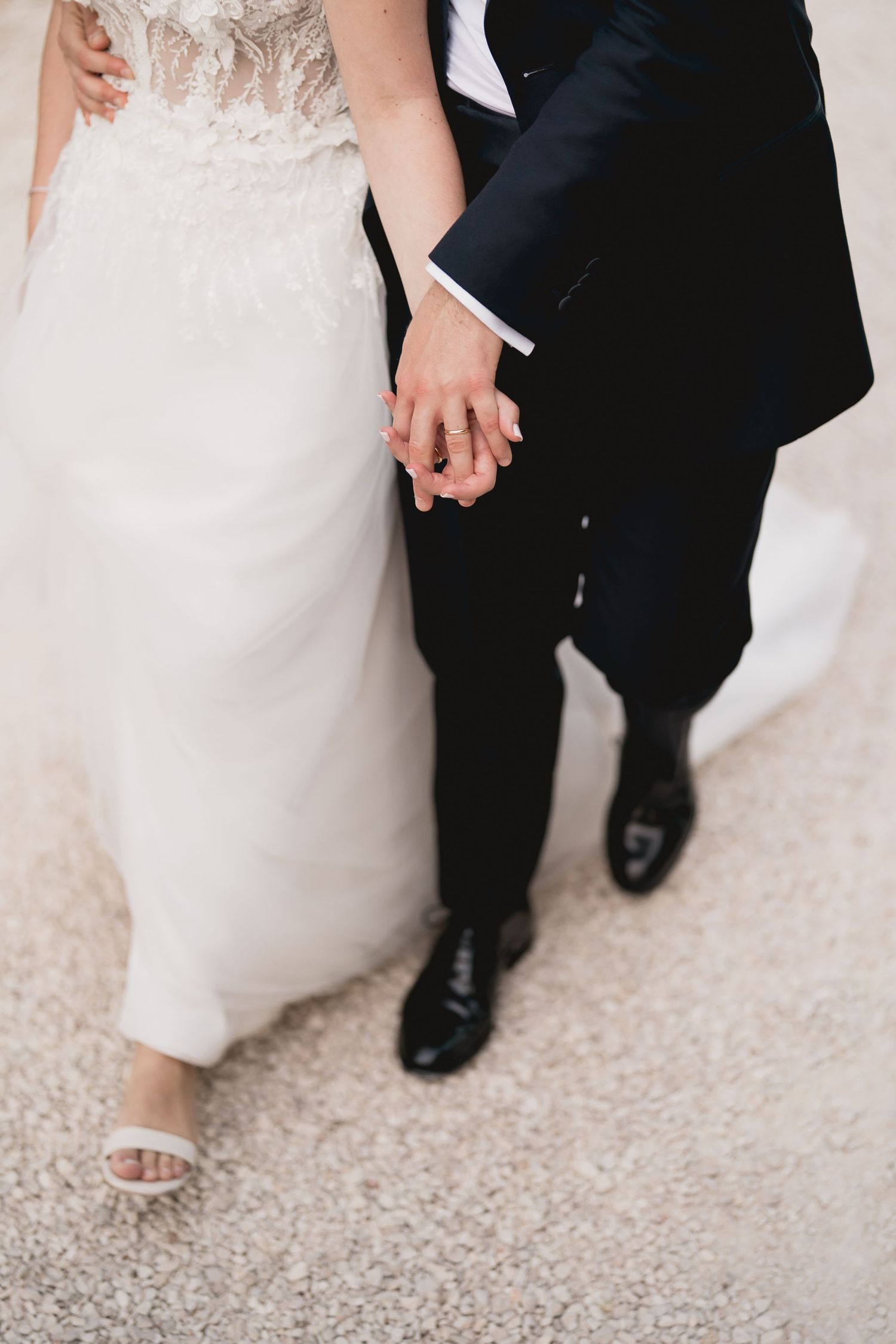 Close-up detail shot of newlyweds holding hands, showing wedding dress lace and classic tuxedo.