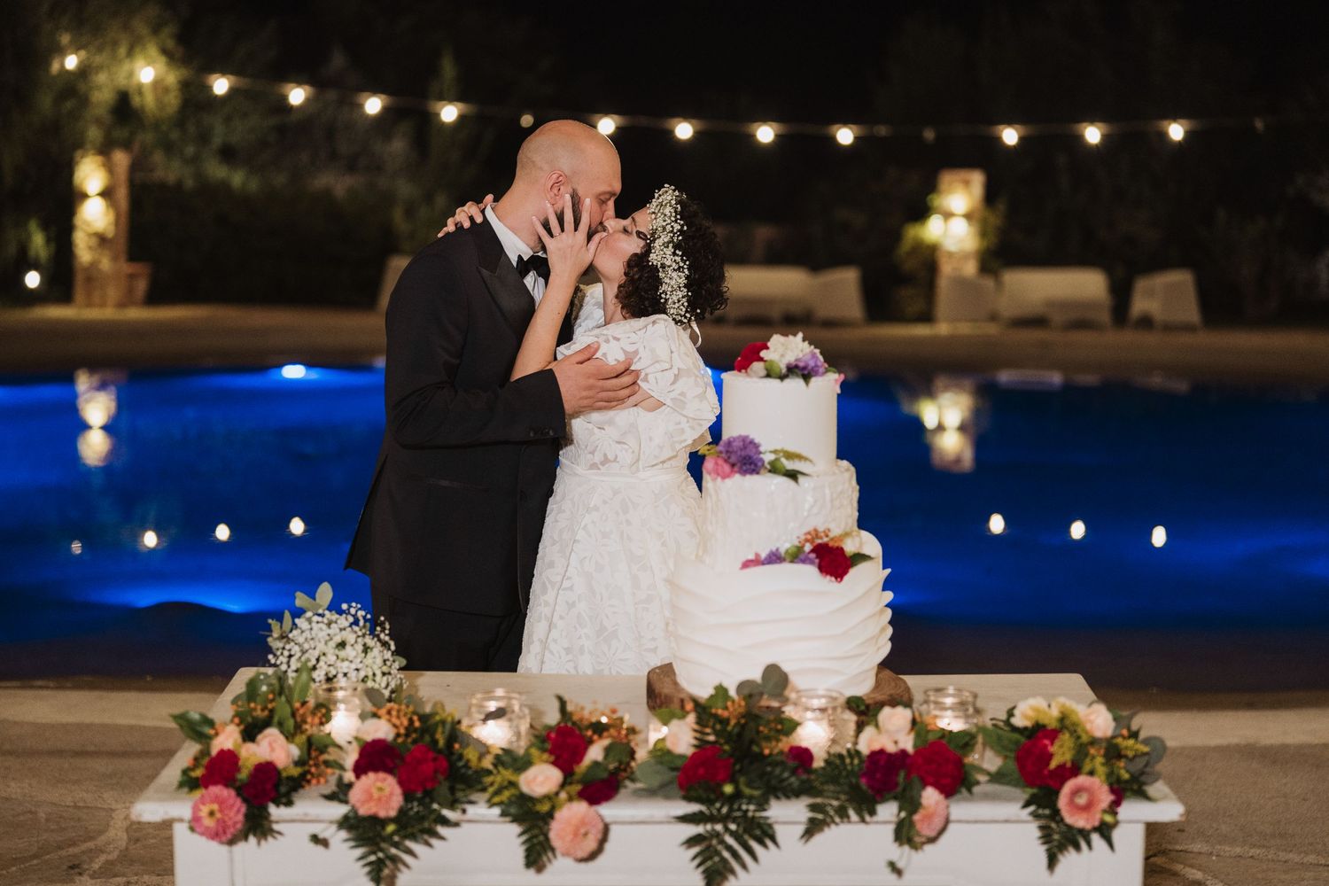 Wedding cake cutting scene by a illuminated pool with red floral decorations.
