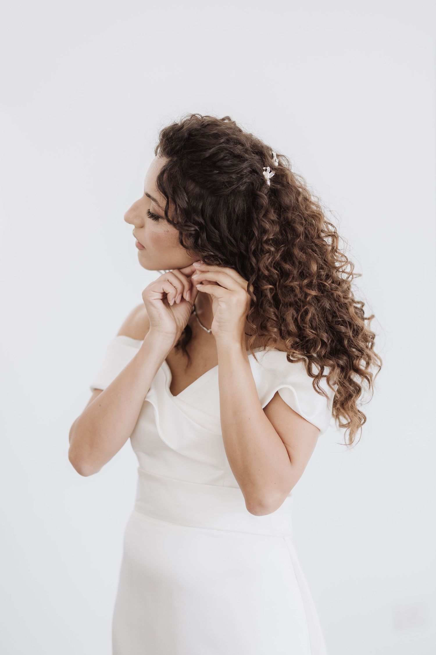 Side view of someone in white dress with curly hair putting on earrings.