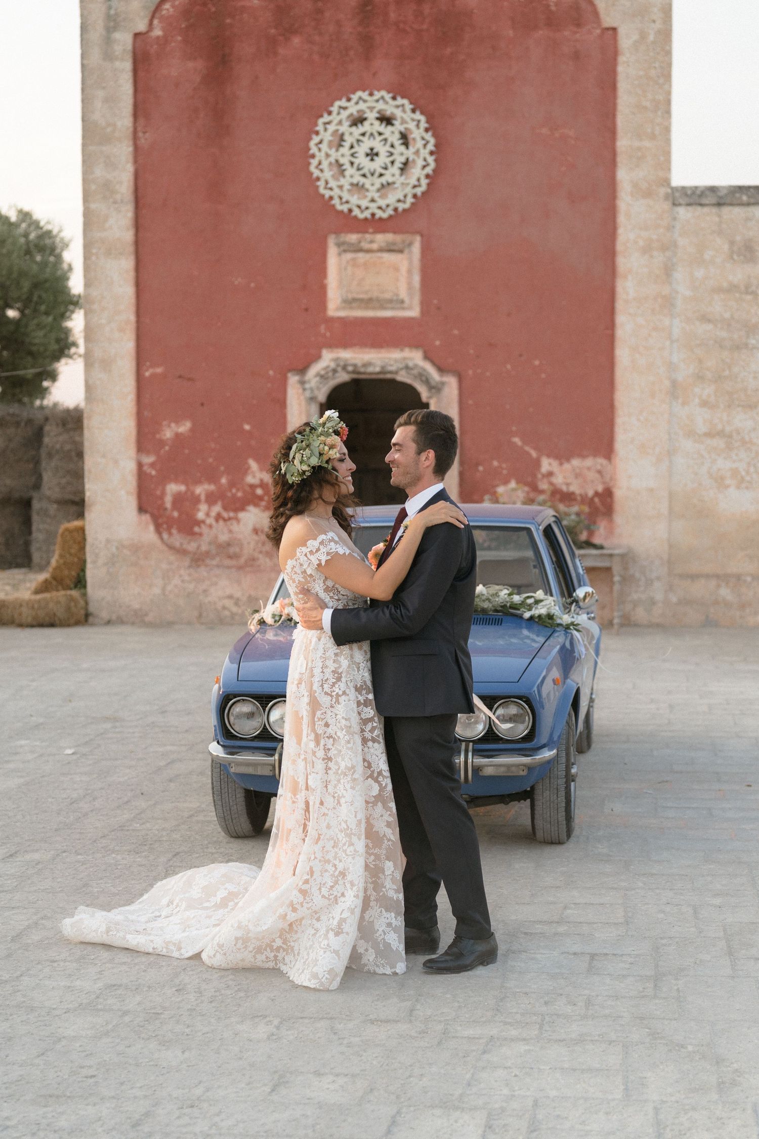 A classic blue vintage car parked against a weathered red wall creates a romantic wedding scene.