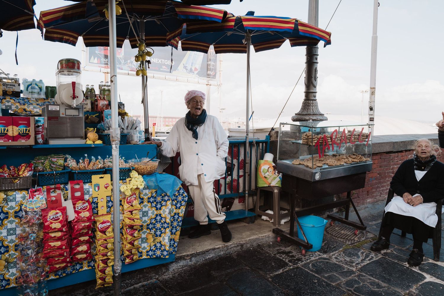Street vendor's cart displays colorful snacks and treats under an umbrella on a city sidewalk.