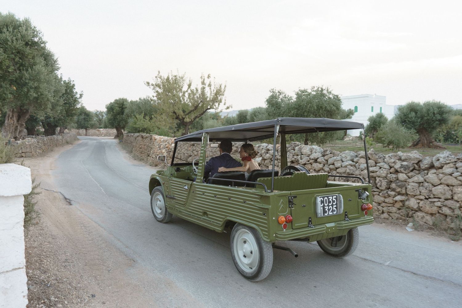 Small green utility vehicle with canopy driving down a narrow rural road bordered by stone walls under an overcast sky.