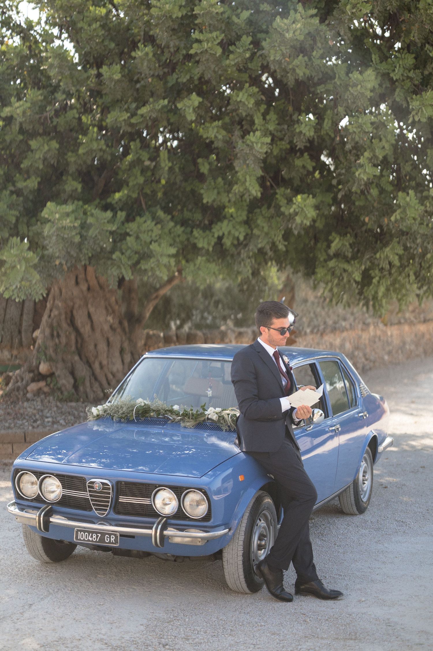 Classic blue Alfa Romeo car parked under trees with someone leaning against it.