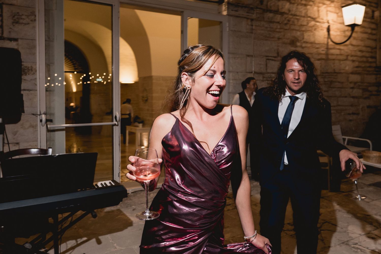 A guest in a burgundy dress enjoys a drink during a festive evening celebration at a rustic indoor venue.