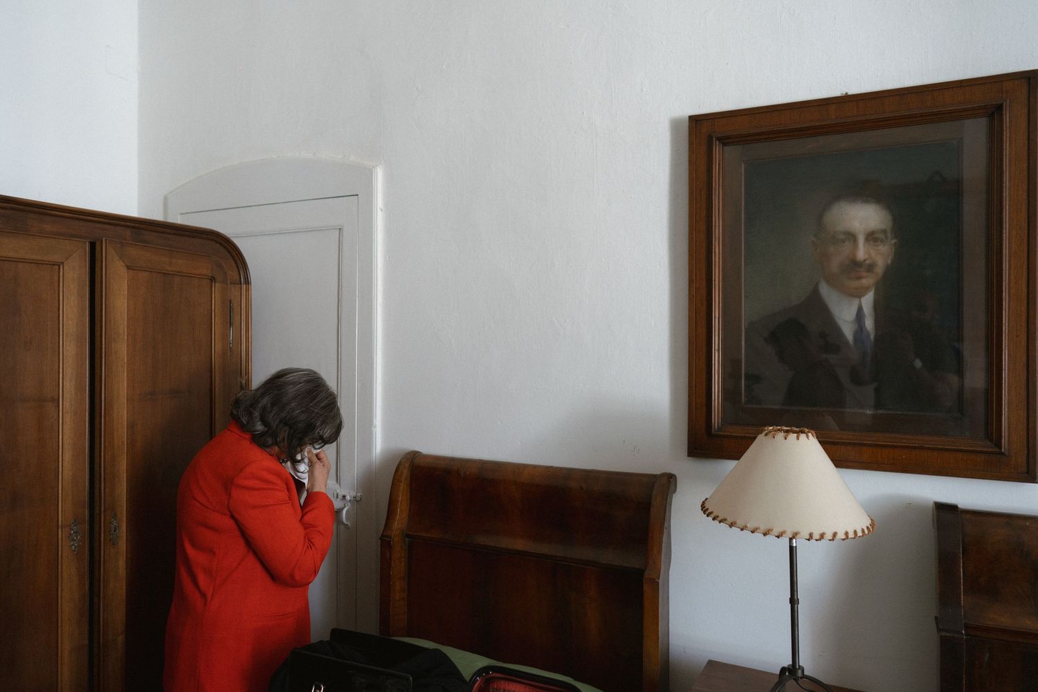 A room with white walls and wooden furniture displays vintage portraits and a table lamp.
