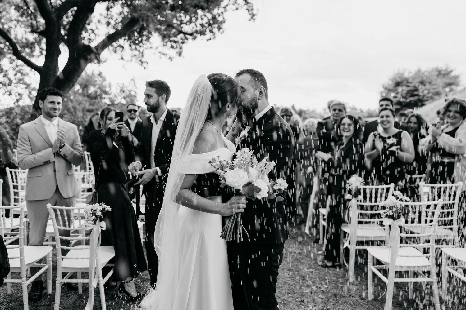 Monochrome image of wedding attendees seated in rows during an outdoor forest ceremony.