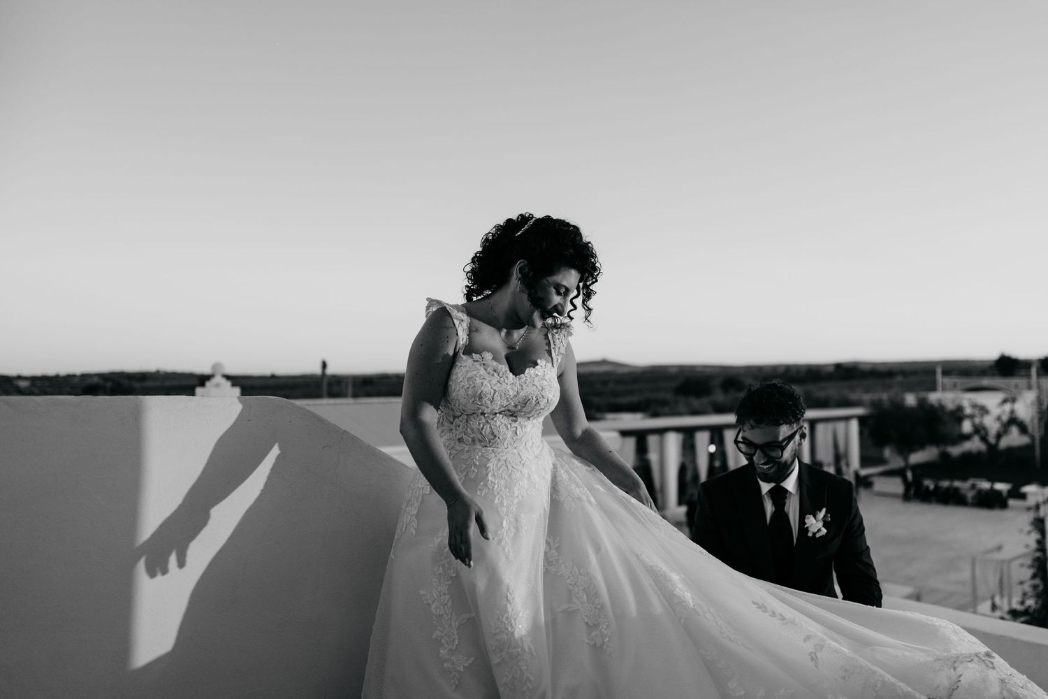 Bride in lace gown with flowing train walks with groom on rooftop in black and white photo.