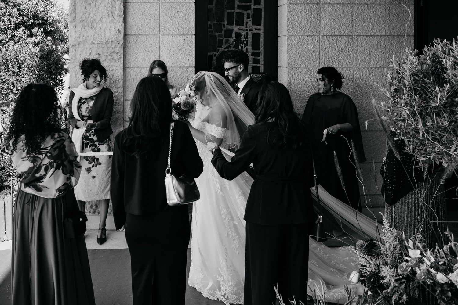 Bride in white gown receives veil adjustment from guests outside church entrance in black and white.