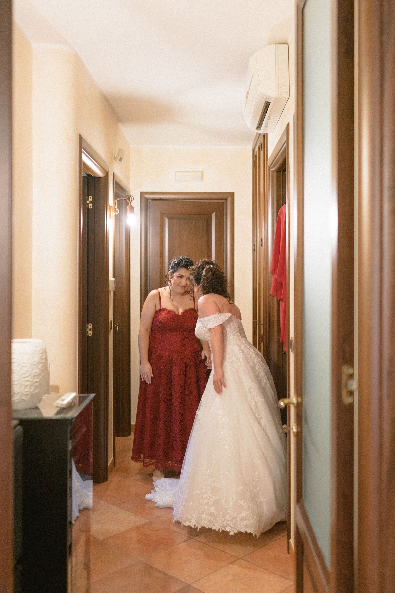 Bride in white gown shares a tender moment with bridesmaid in red lace dress in a warmly lit hallway.