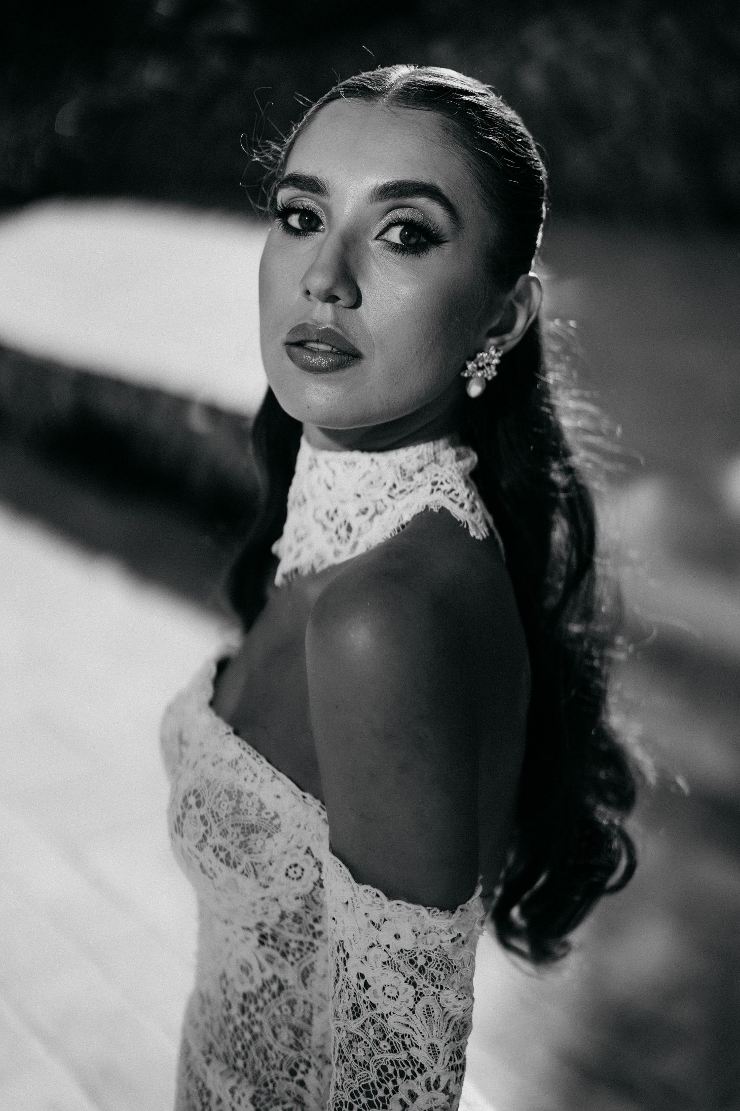 Black and white close-up portrait of a bride in a lace off-shoulder wedding dress with a high collar and sparkling earrings.
