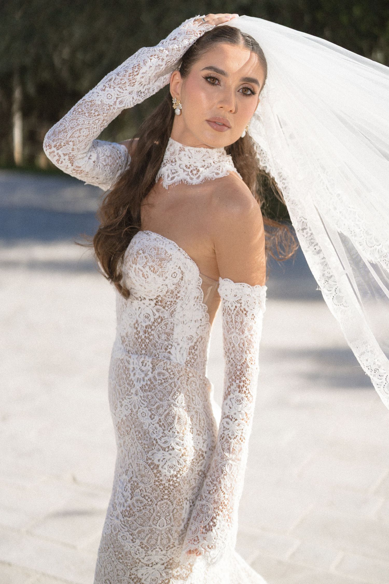 Bride in an off-the-shoulder lace wedding gown with long lace gloves, high lace collar and a flowing veil poses outdoors.