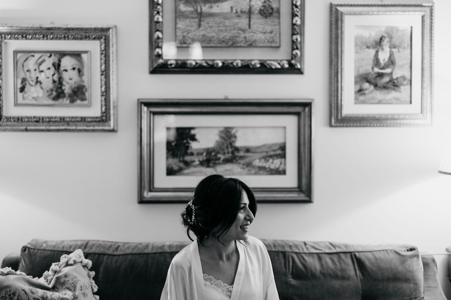 A bride in a white robe sits on a sofa smiling, with framed artwork displayed on the wall behind her.