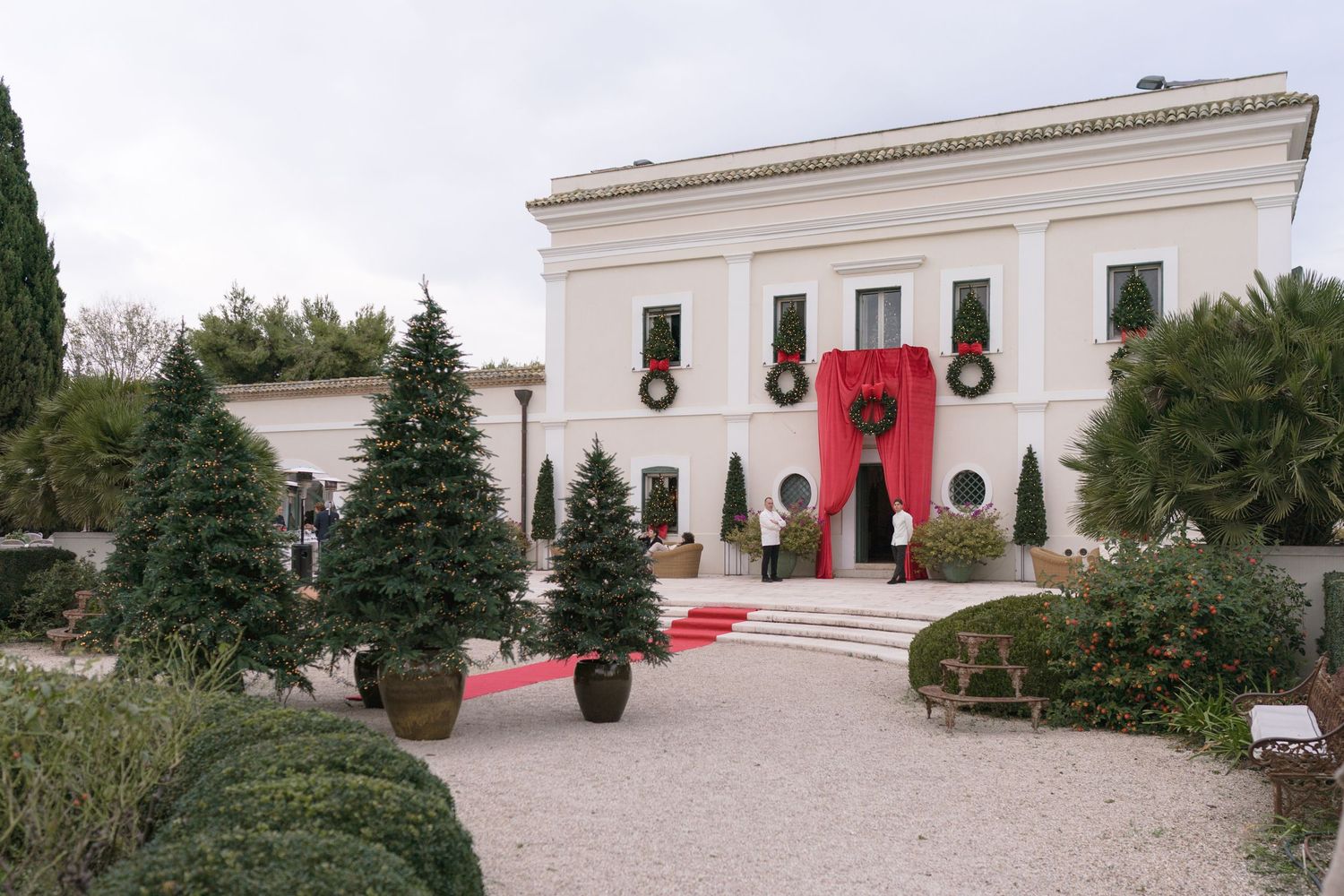 Elegant white villa decorated with red holiday wreaths and garlands surrounded by manicured evergreen trees.