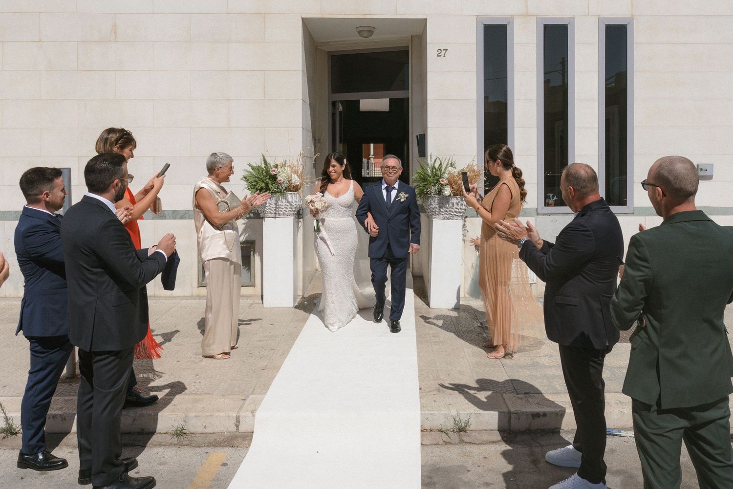 Wedding party and guests gathered outside white stone building entrance.