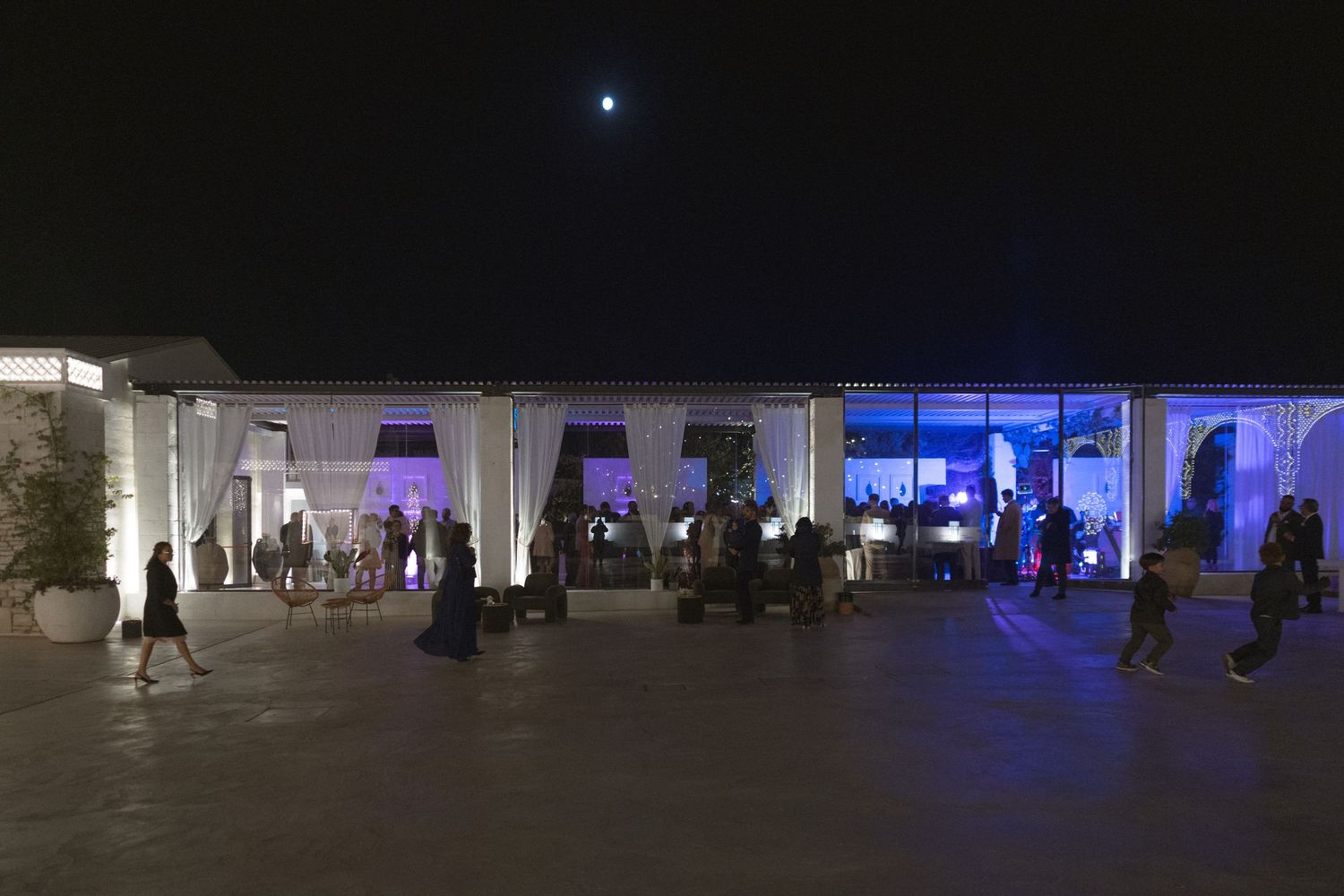 Outdoor evening wedding reception venue illuminated with blue lights under a full moon with guests mingling.