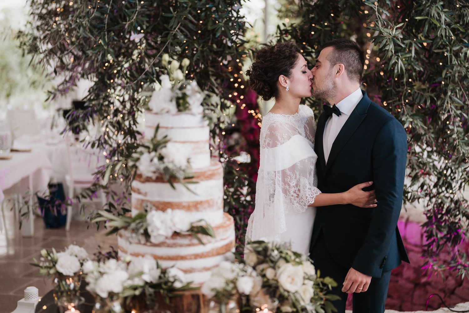 Romantic wedding scene with floral decorations and a multi-tiered cake under hanging greenery and lights.