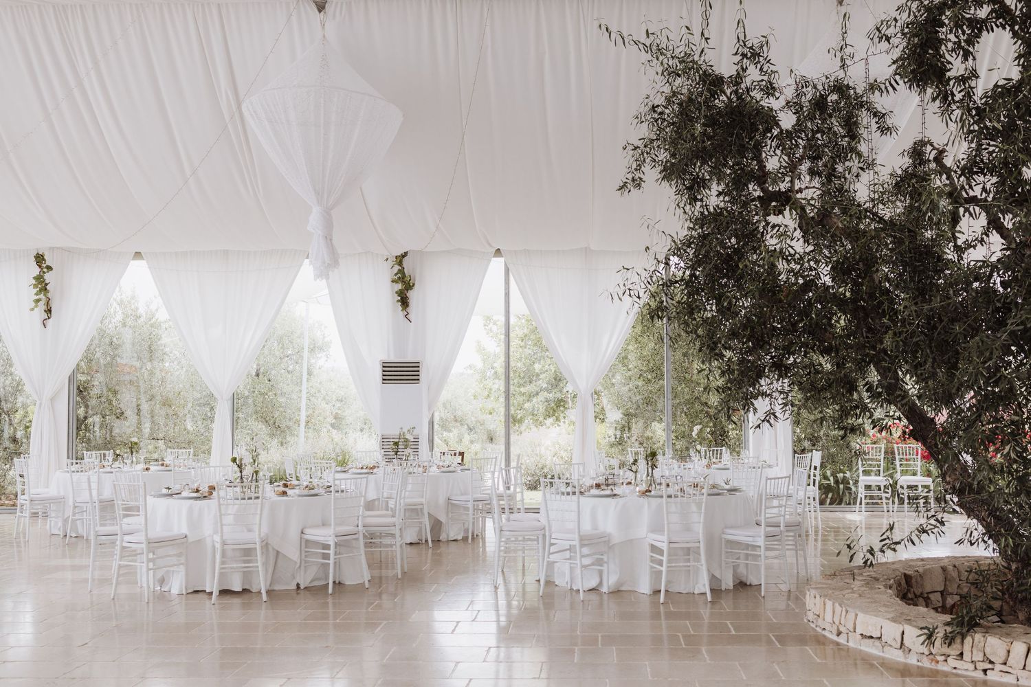 Elegant white wedding reception venue with draped ceilings, chandeliers and round tables set for dining.