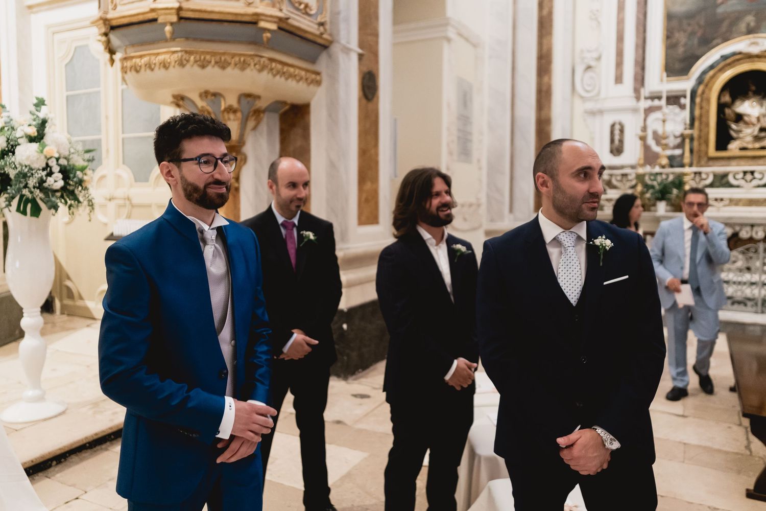 Wedding attendants stand in formal attire inside ornate church interior.
