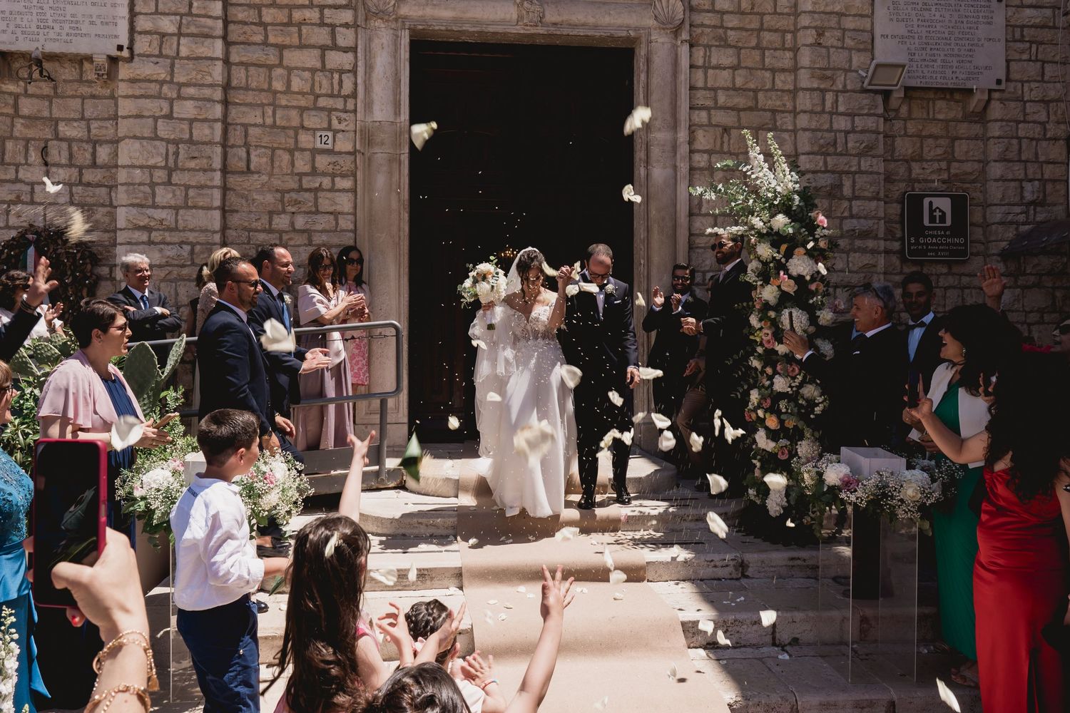 Wedding guests shower rice on the newlyweds as they exit a stone church on a sunny day.