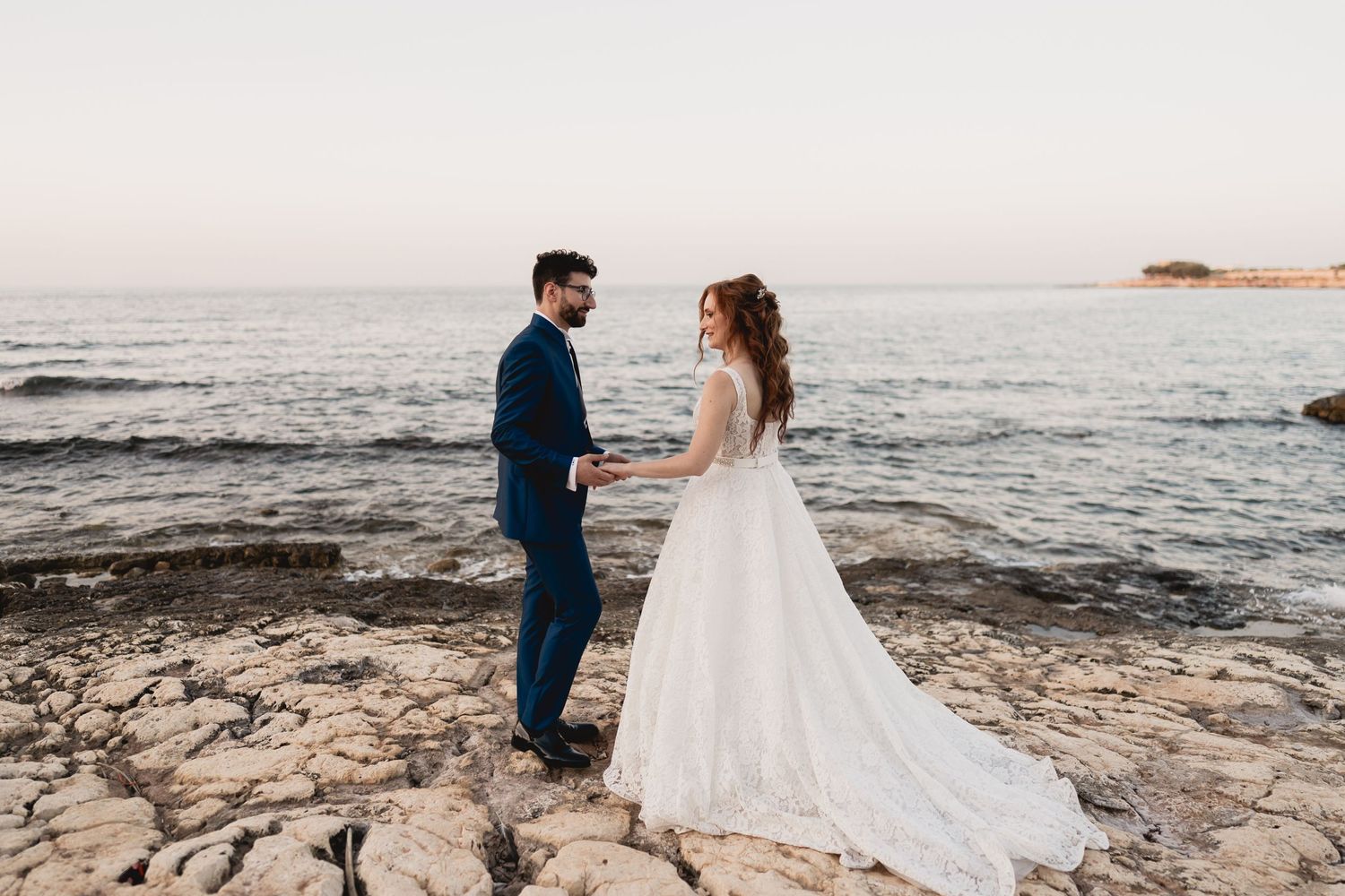 A romantic wedding photo captured at sunset on rocky coastal cliffs overlooking the ocean.