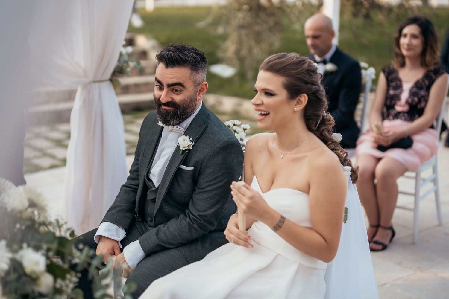 Wedding ceremony scene with couple seated under white draped decor outdoors.