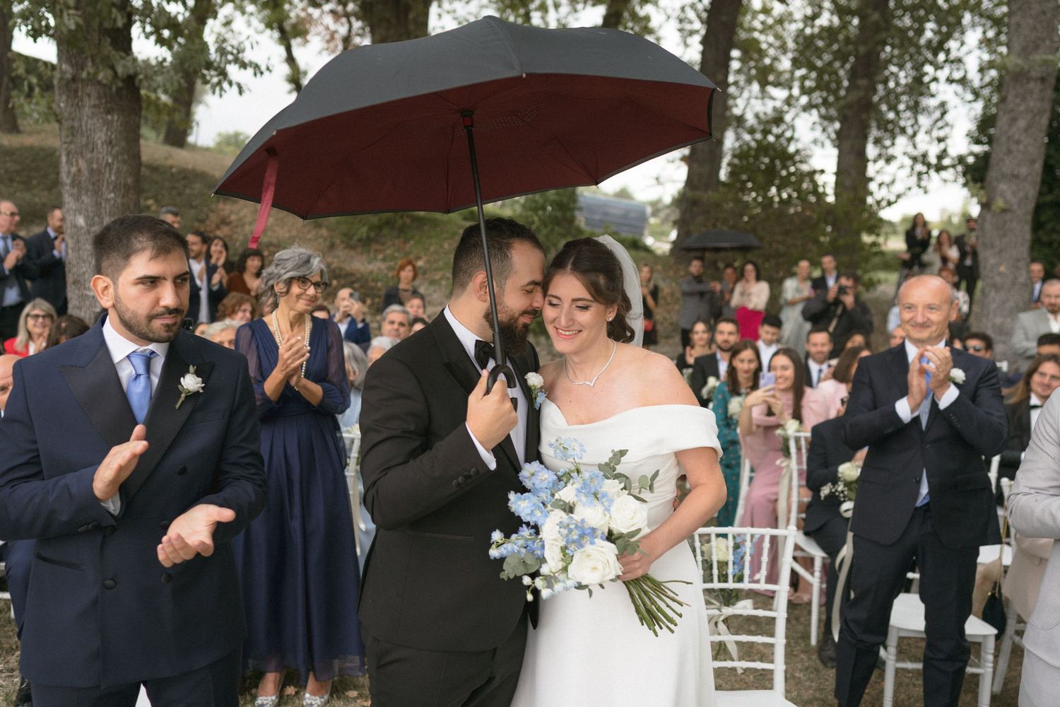 Wedding couple shares an intimate moment under a dark umbrella while guests celebrate in the background.